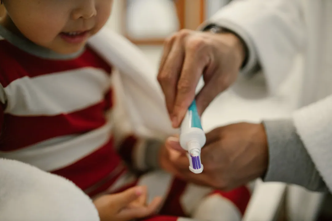 An adult's hands squeeze toothpaste onto a young child's toothbrush, with the child's face cropped below the eyes