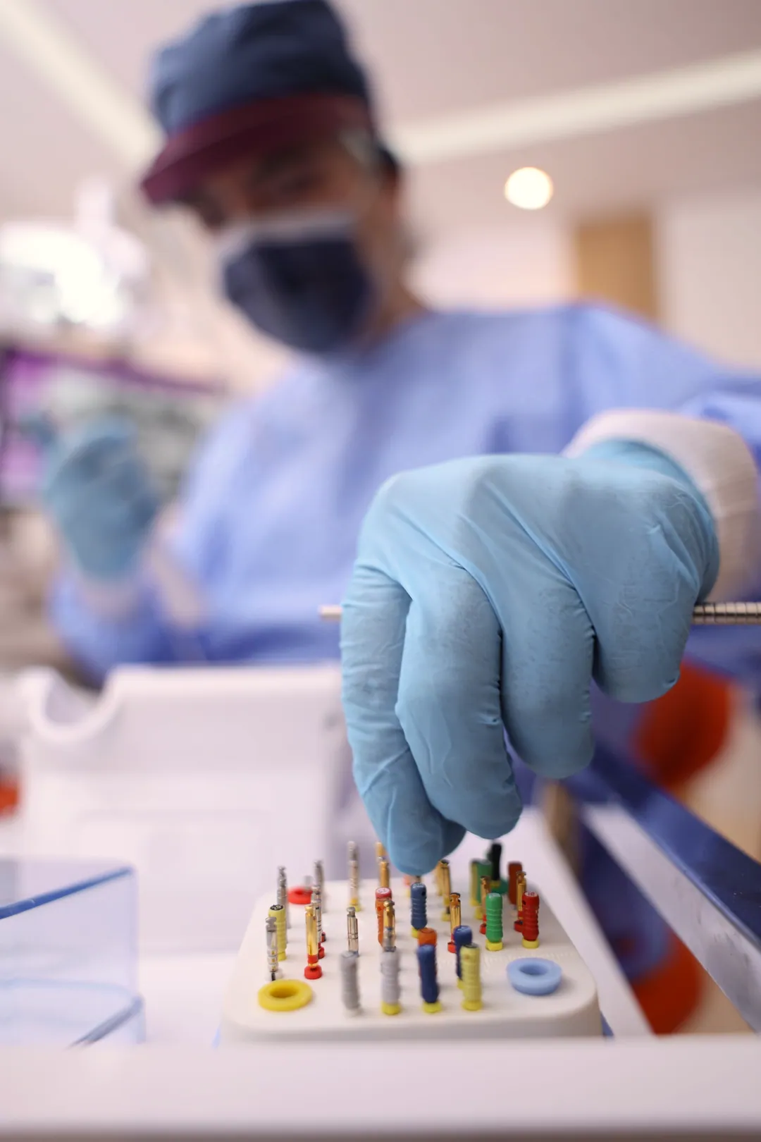 A gloved hand picks an endodontic file from a stand, with a masked clinician blurred behind