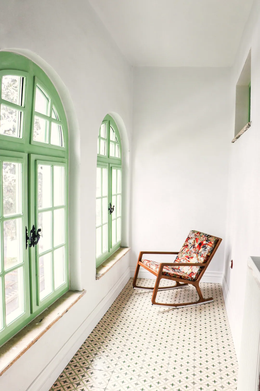 A sunlit white corridor with green-framed arched windows and a single armchair on a patterned tile floor
