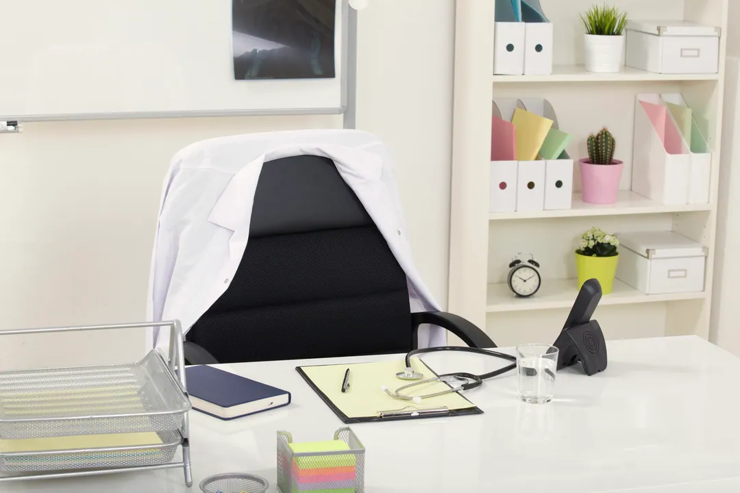 A white coat draped over a desk chair, with a stethoscope, notebook and files on the desk