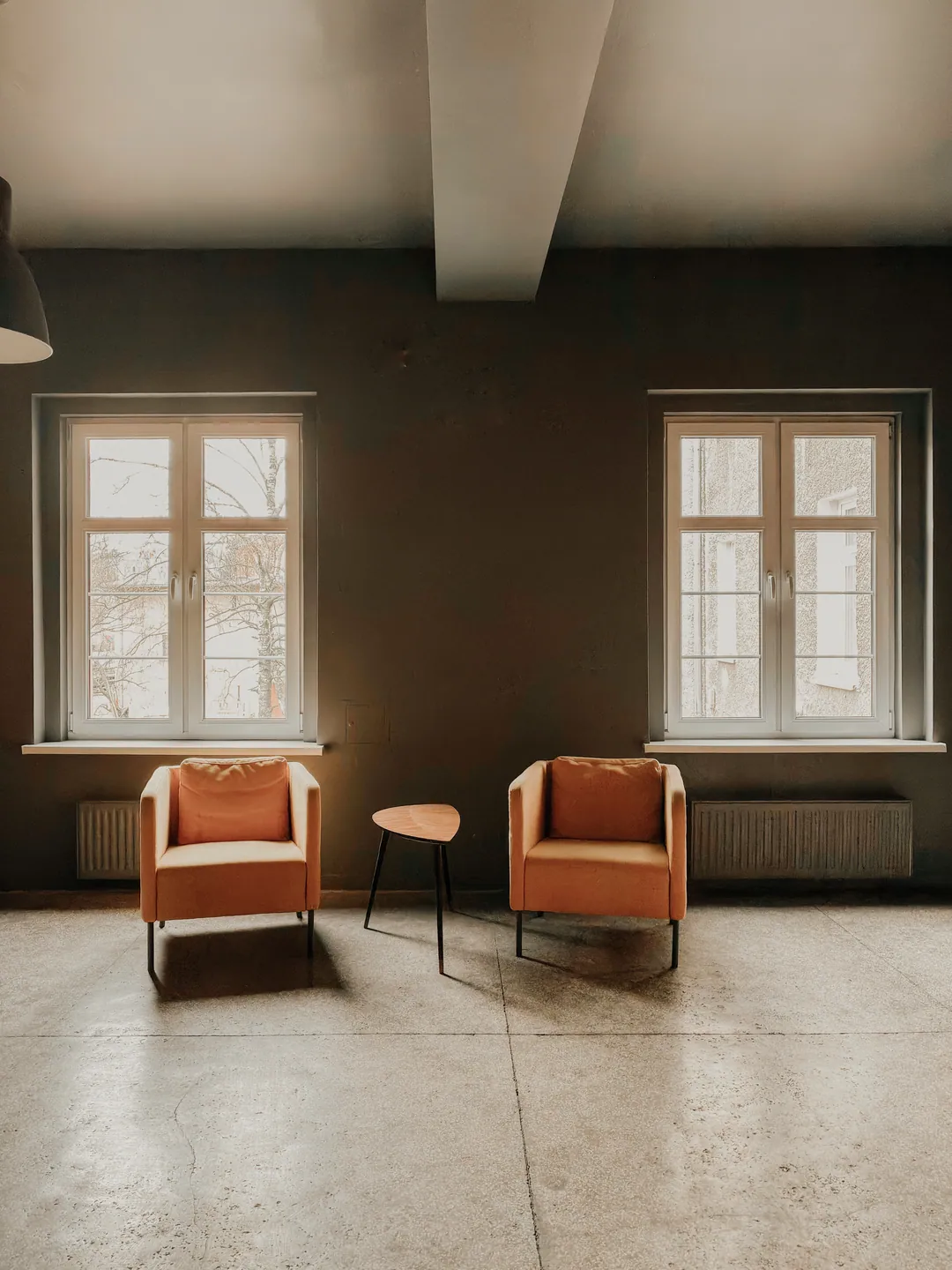 Two terracotta armchairs and a small table between two tall windows in a calm, sunlit room