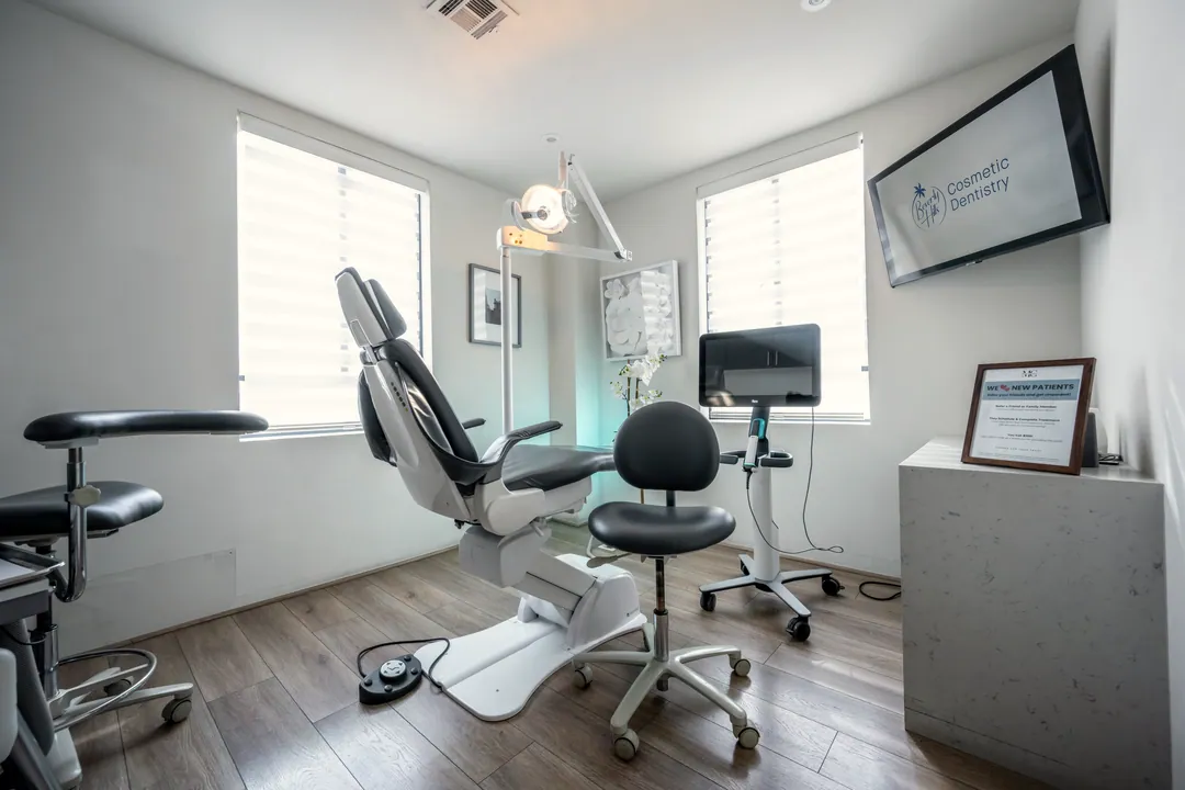 An empty treatment room with a chair, stool, monitor and two windows