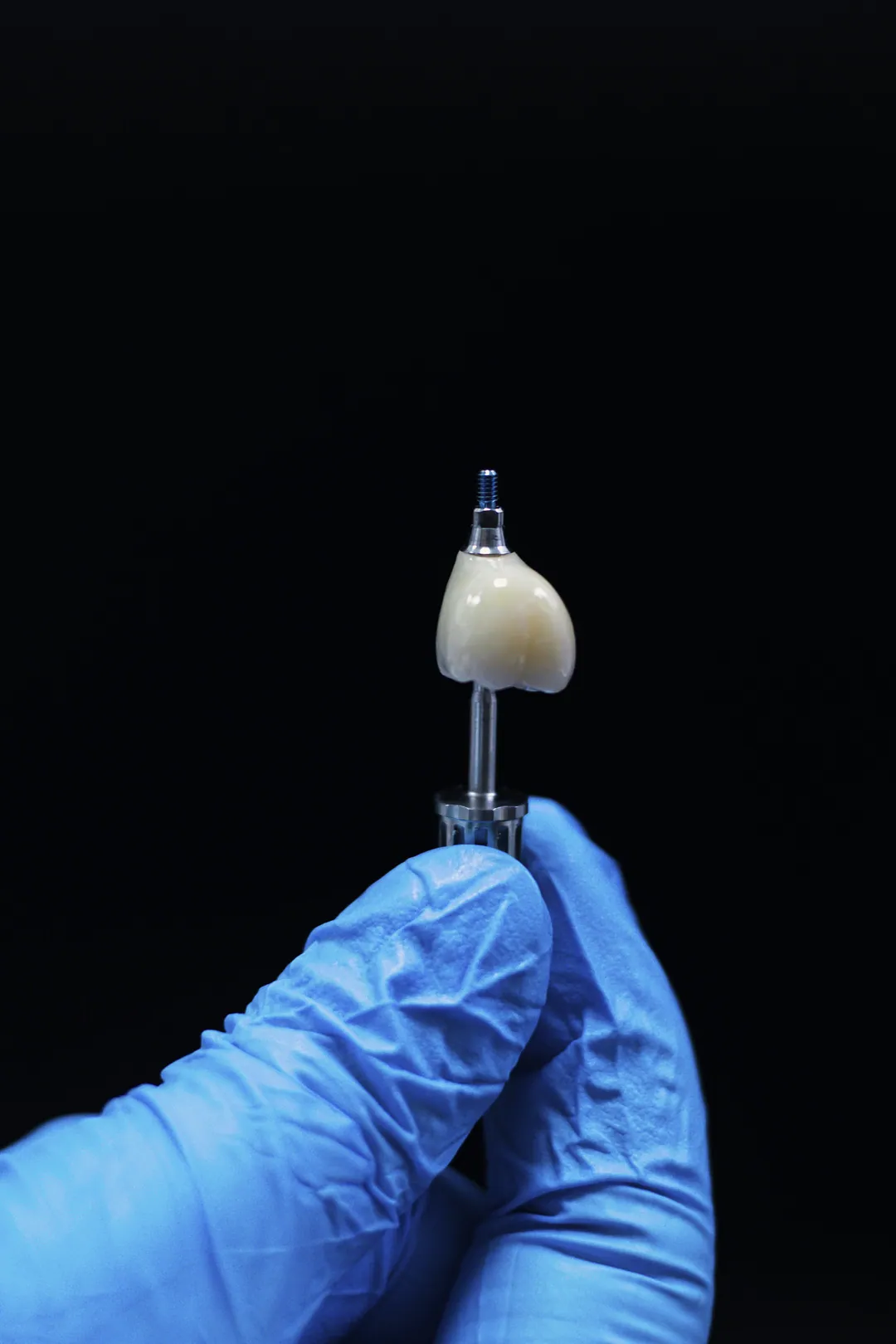 A blue-gloved hand holds an implant with a crown on a black background