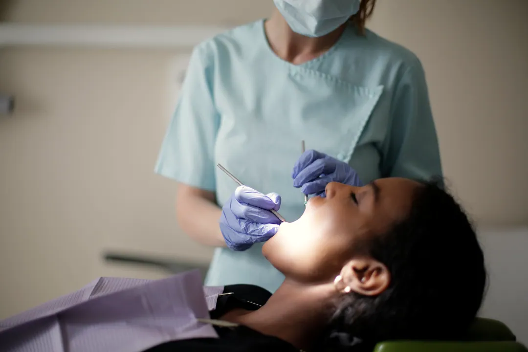 A clinician in pale-blue scrubs examines a reclining patient with a mirror and probe under the lamp