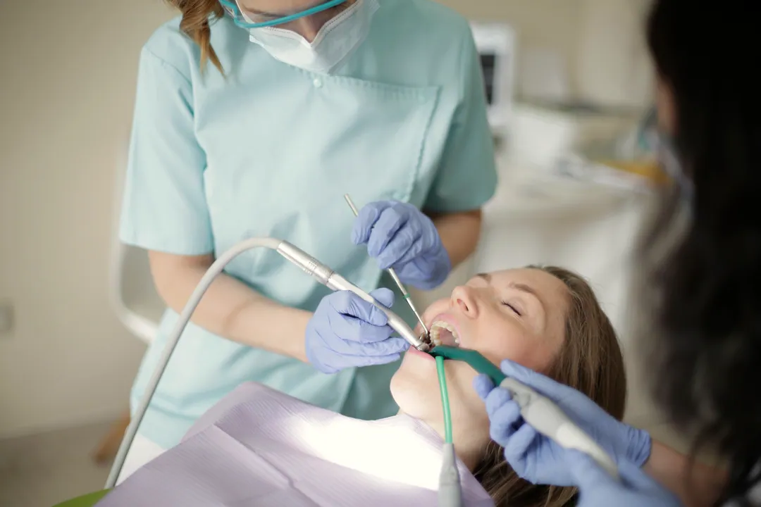 A clinician scales a patient's teeth with an ultrasonic scaler while an assistant holds suction