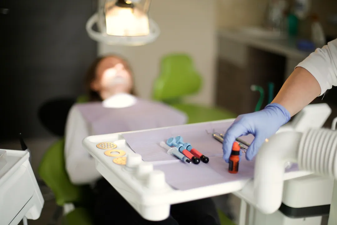 A gloved hand reaches for material on the instrument tray, with a patient blurred in the chair