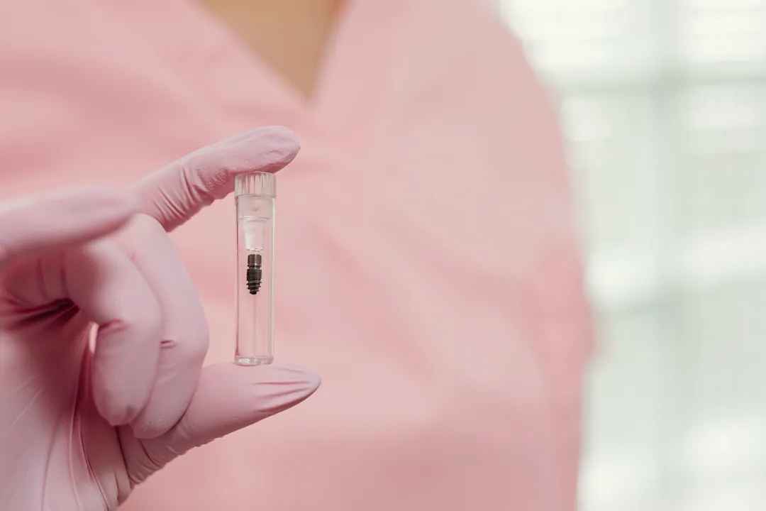 A pink-gloved hand holds a small sterile vial containing a dental implant