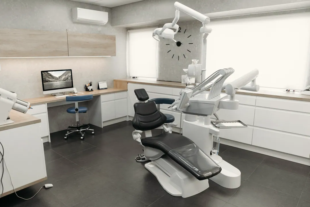 An empty modern treatment room with a dark dental chair, cabinets, a desk and a wall clock in daylight