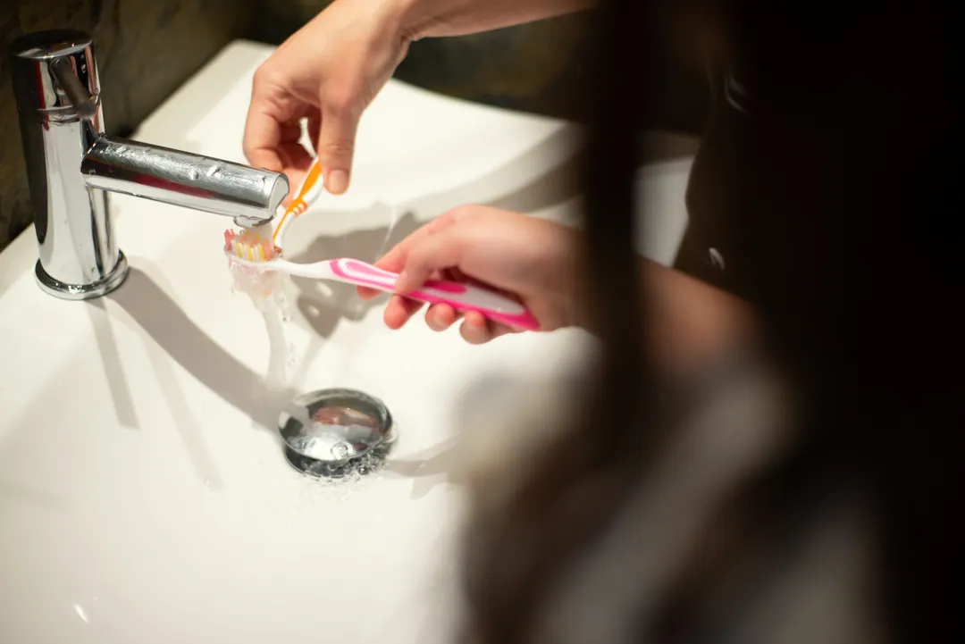 Two hands rinse toothbrushes under a tap at a white basin