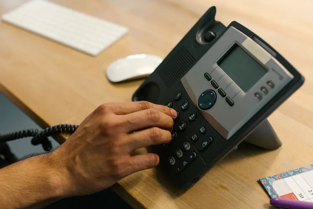 A hand dials a desk telephone on a light wood desk next to a keyboard