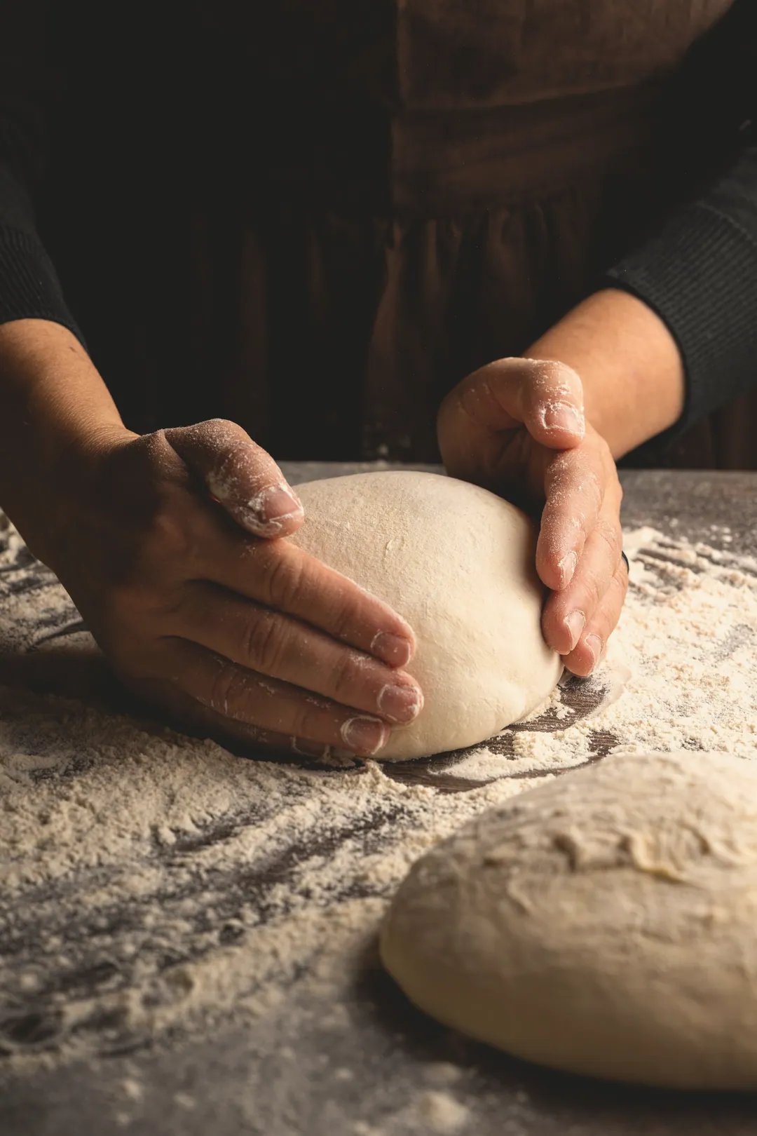 A baker's hands rounding dough on a floured bench