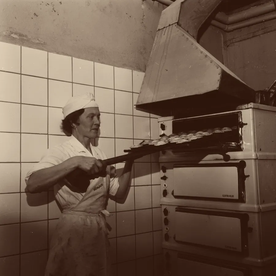 Black and white photograph: a woman baker in whites slides a tray of buns into a deck oven with a long peel