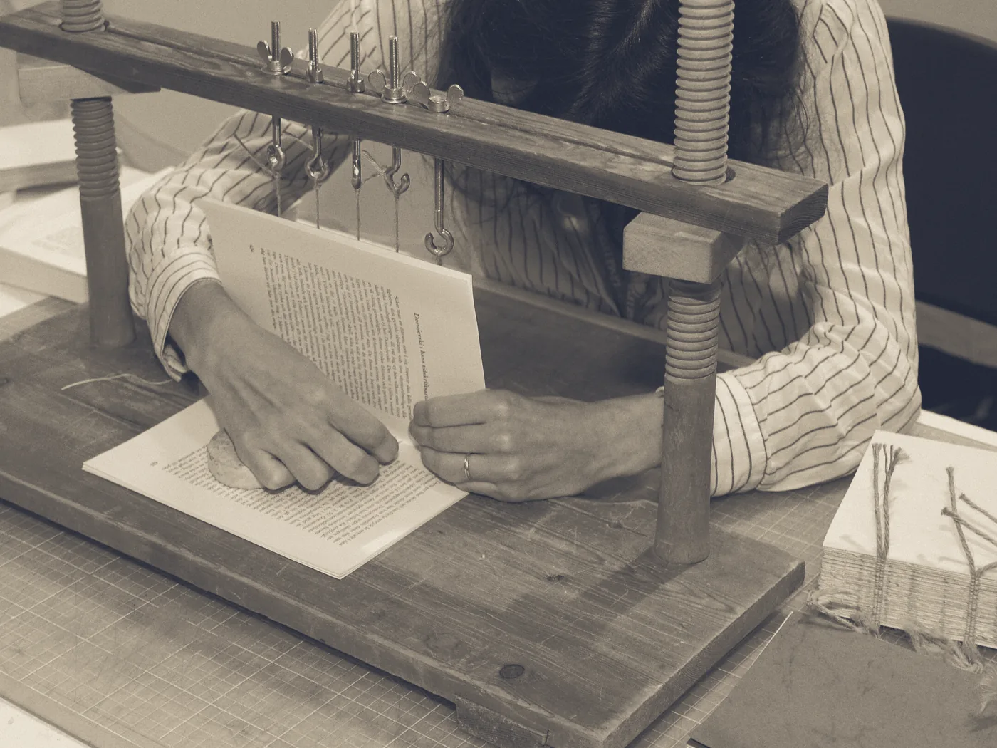 Hands at a sewing frame, passing the thread through the fold of a signature, a finished book block beside it