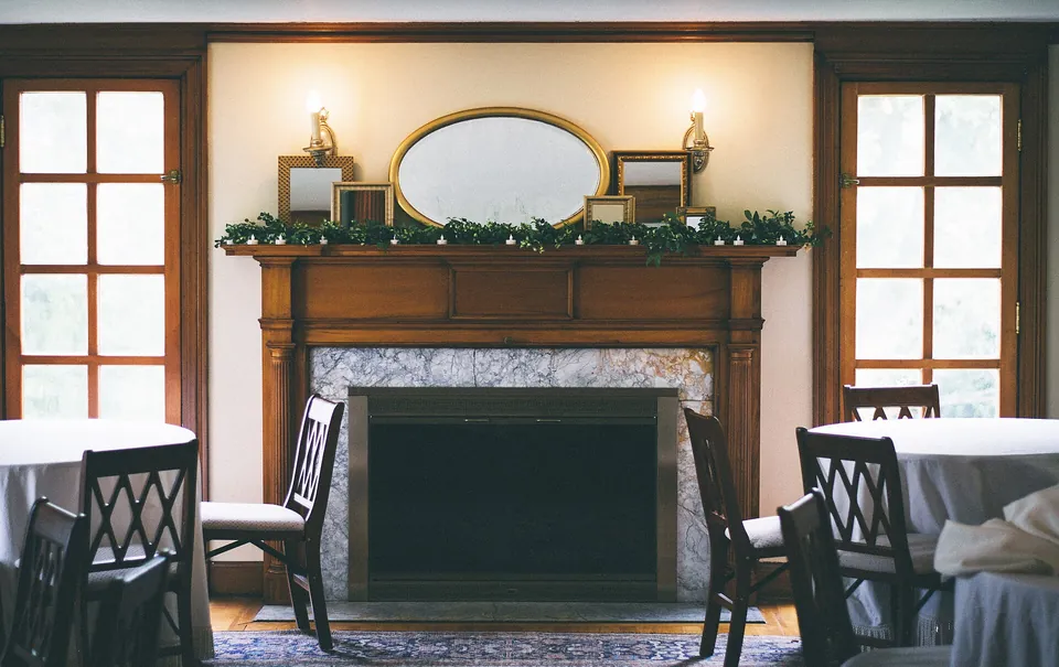 A dining room set for lunch: round tables in white cloths, a fireplace with greenery and two wall lamps