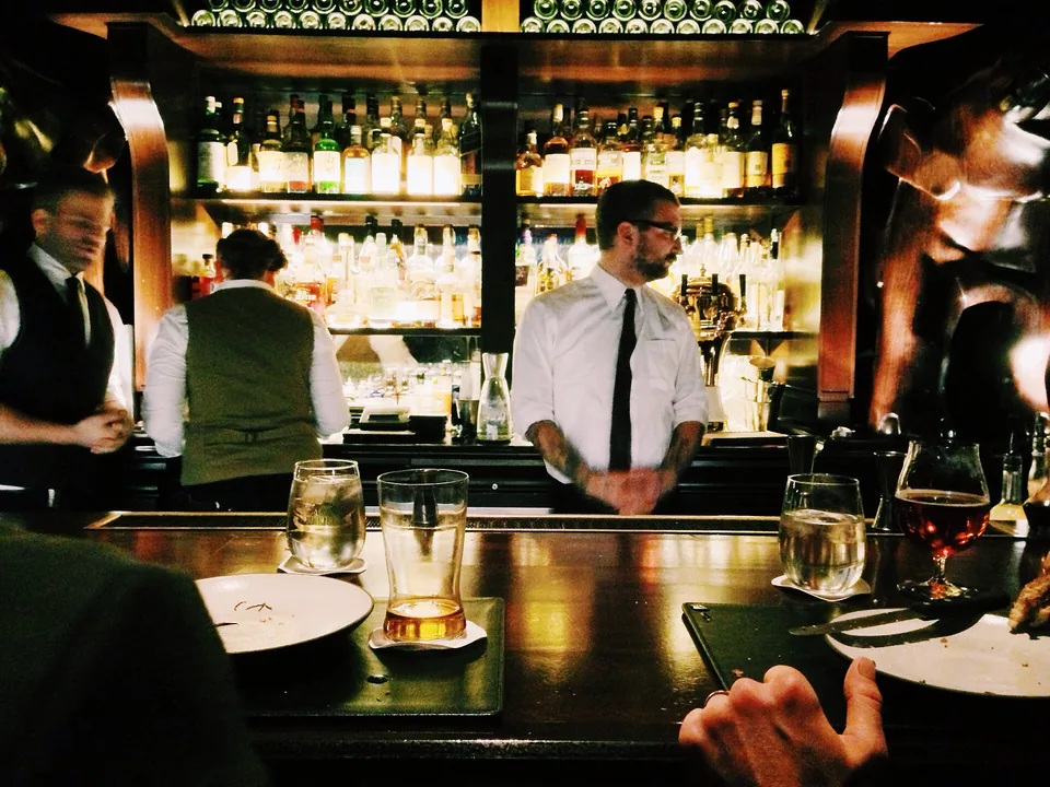 A full counter at night: bartenders in shirtsleeves, back-lit shelves of bottles, glasses and plates on the bar
