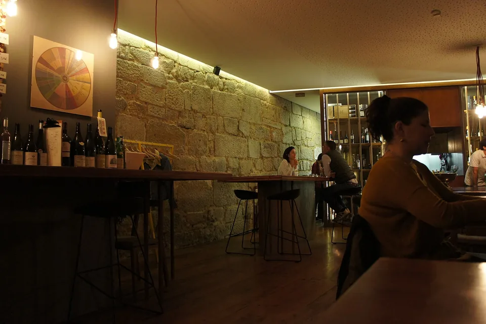 A wine bar with a sandstone wall washed by a strip light, bottles on shelves, a guest at a window table