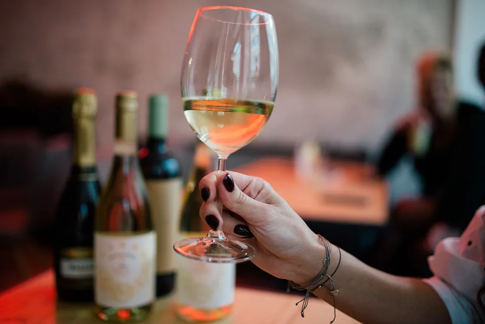 A glass of white wine raised in front of three bottles, a lively bar behind