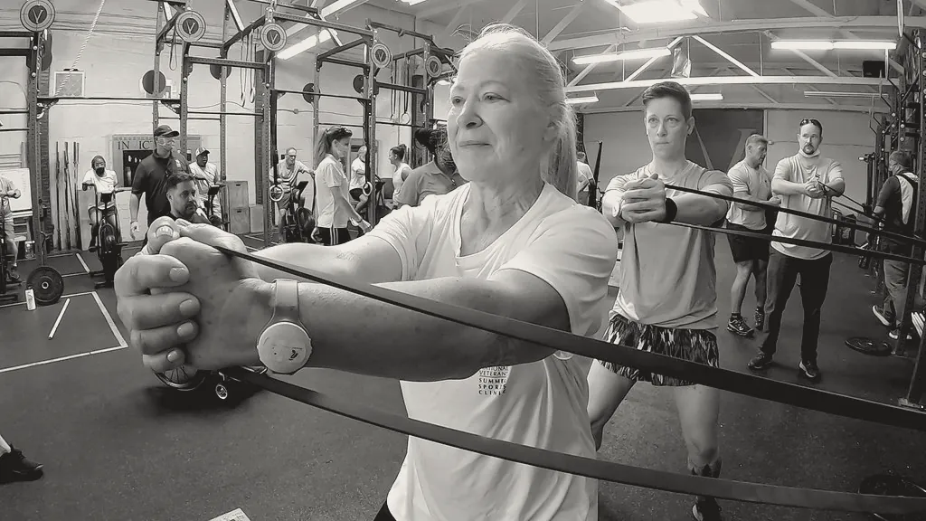 A woman in her sixties stretching a band at shoulder height, a coach and a small group behind her.