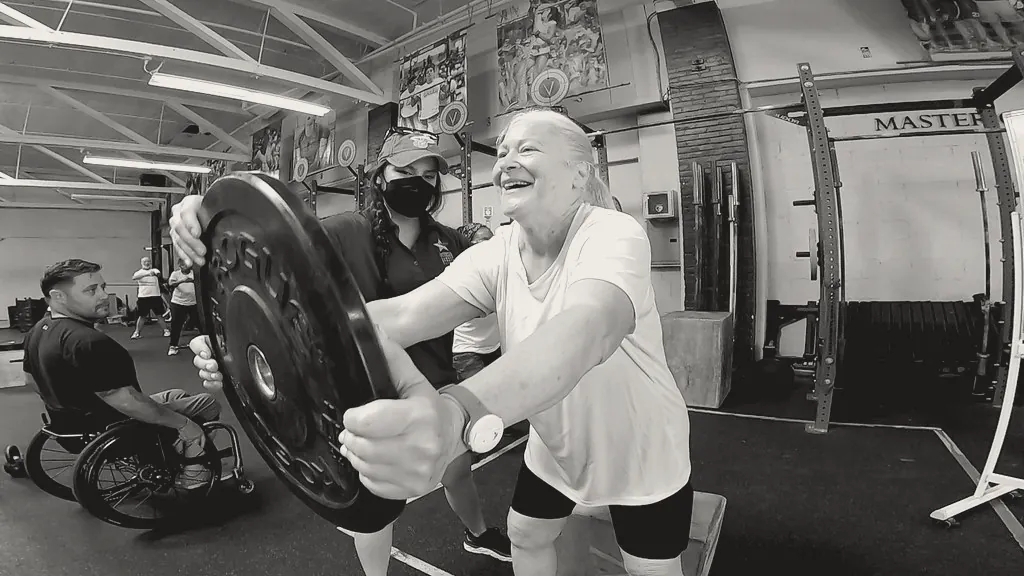 An older woman laughing as she presses a plate forward, a coach beside her.