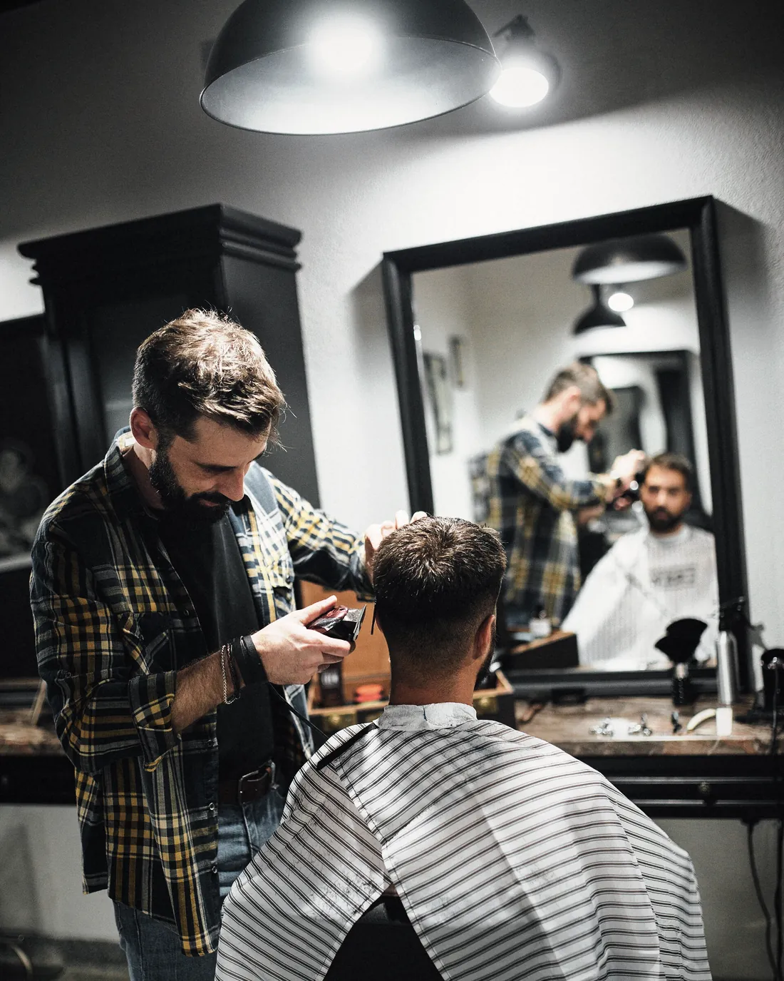 A barber at work in a shop with tall mirrors and hanging lamps