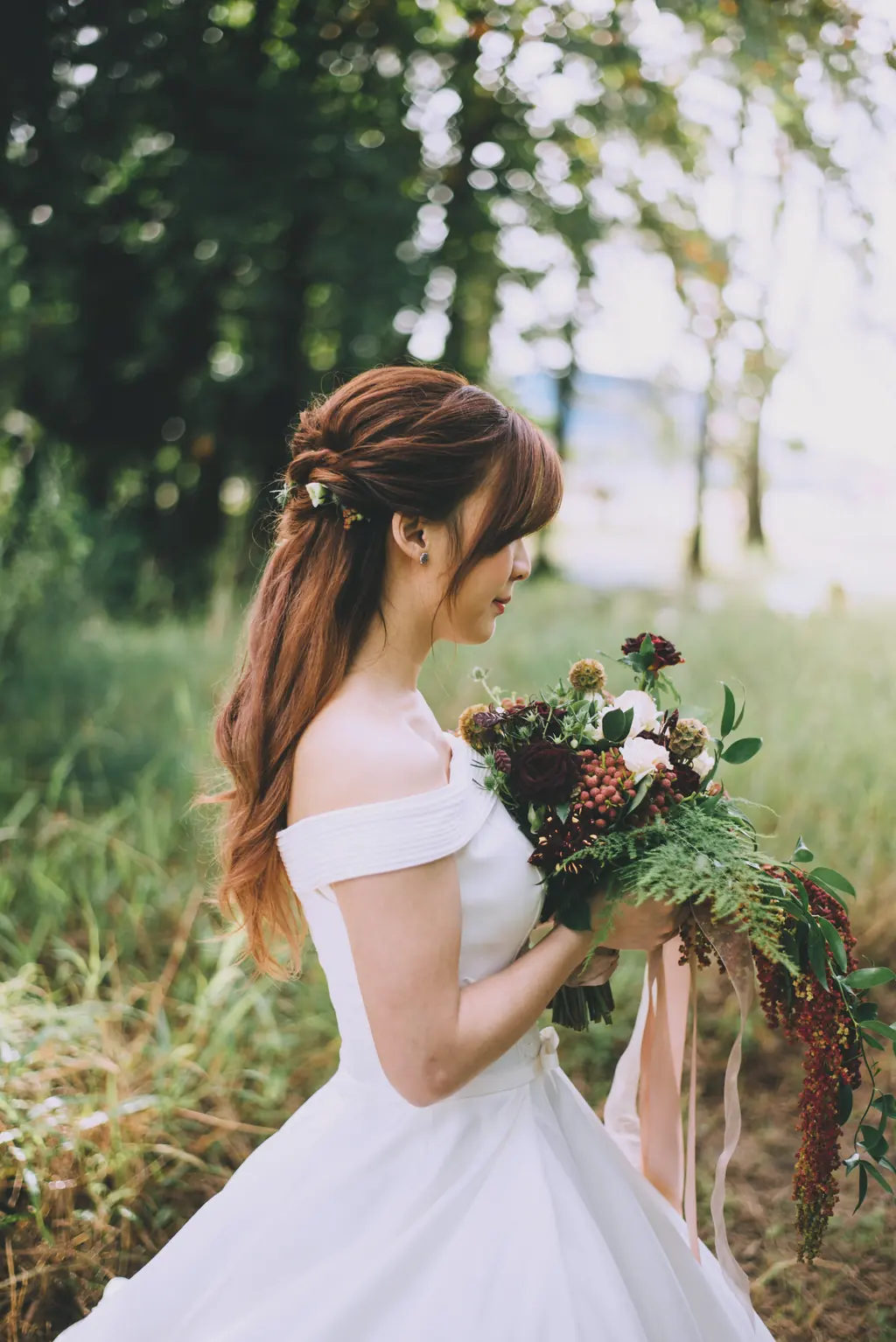 A bride in a meadow holding a bouquet of burgundy flowers and trailing green