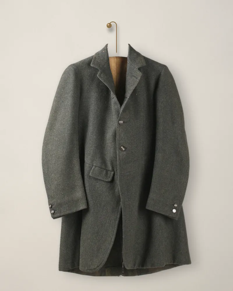 A long single-breasted jacket in dark green loden with a flap pocket, hanging from a brass peg