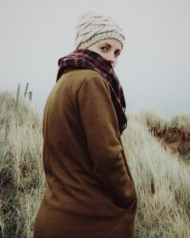 A woman in a long brown tweed coat and a dark scarf, looking back over her shoulder in coastal dunes