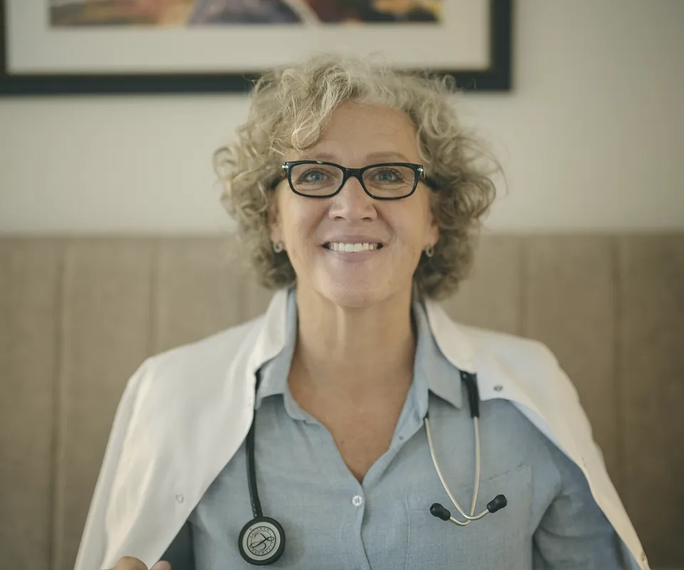 A smiling doctor with grey curly hair and glasses sitting in her office.