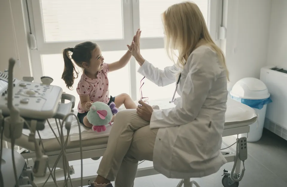 A doctor gives a young girl a high five as she sits on the examination couch, a soft toy on her lap.
