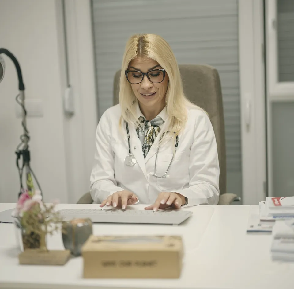 A doctor typing at her desk, a small vase of flowers beside the screen.