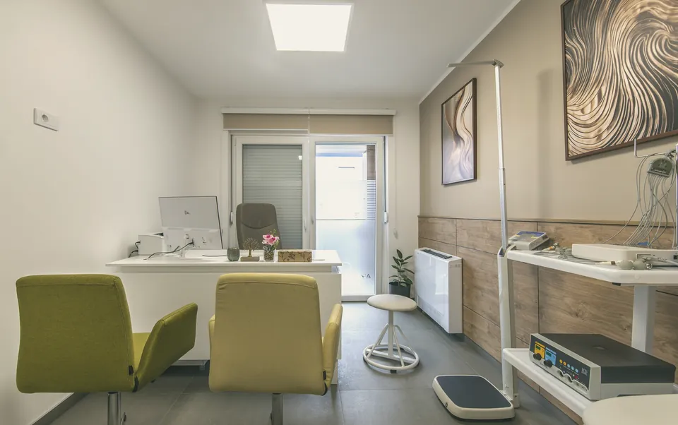 A consulting room in daylight: two olive-green chairs facing the doctor's desk, a window behind.