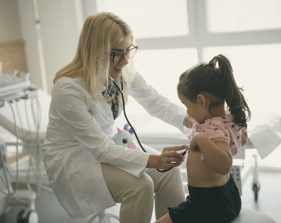 A doctor listening to a little girl's back with a stethoscope while the girl holds up her shirt.