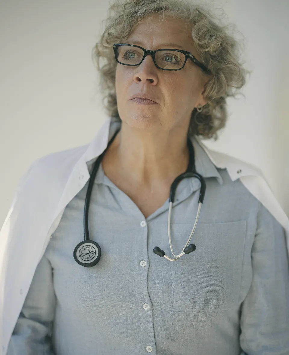 A doctor with grey curly hair and glasses, stethoscope round her neck, standing in her consulting room.