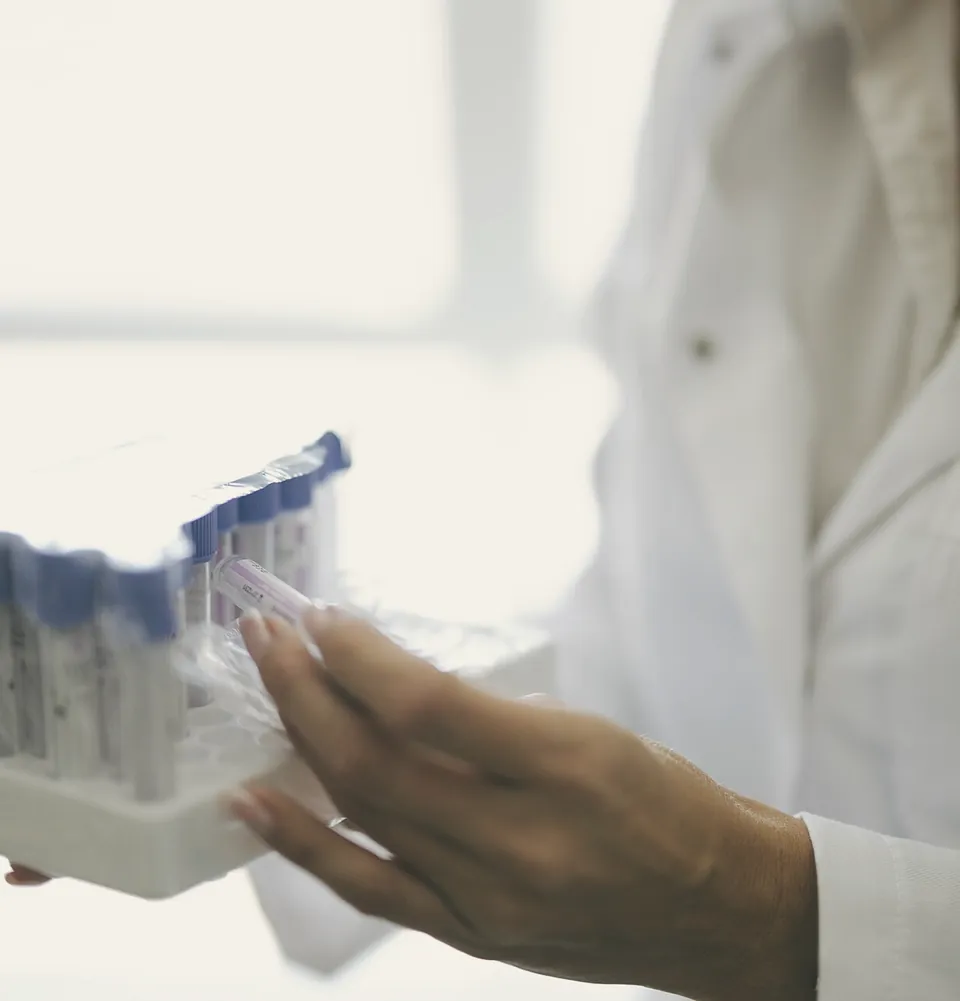 Hands in a white coat holding a tray of labelled blood tubes in front of a bright window.