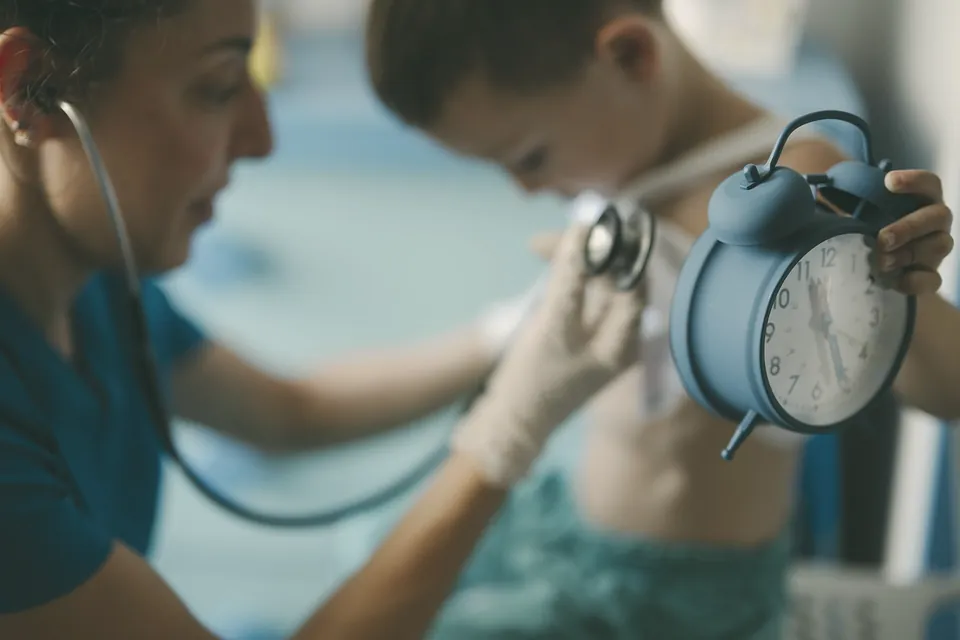 A nurse listening to a boy's chest while he holds up a blue alarm clock.