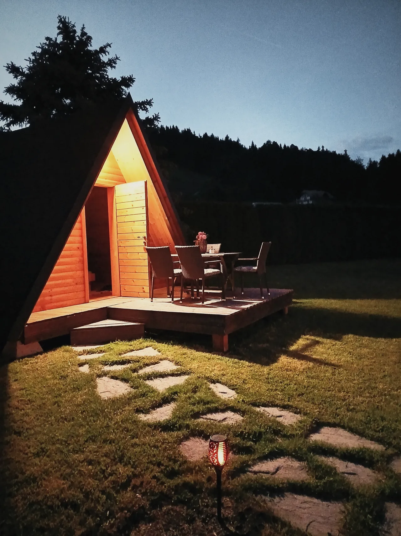 A small A-frame timber cabin lit from inside at dusk, a stone path up to its deck