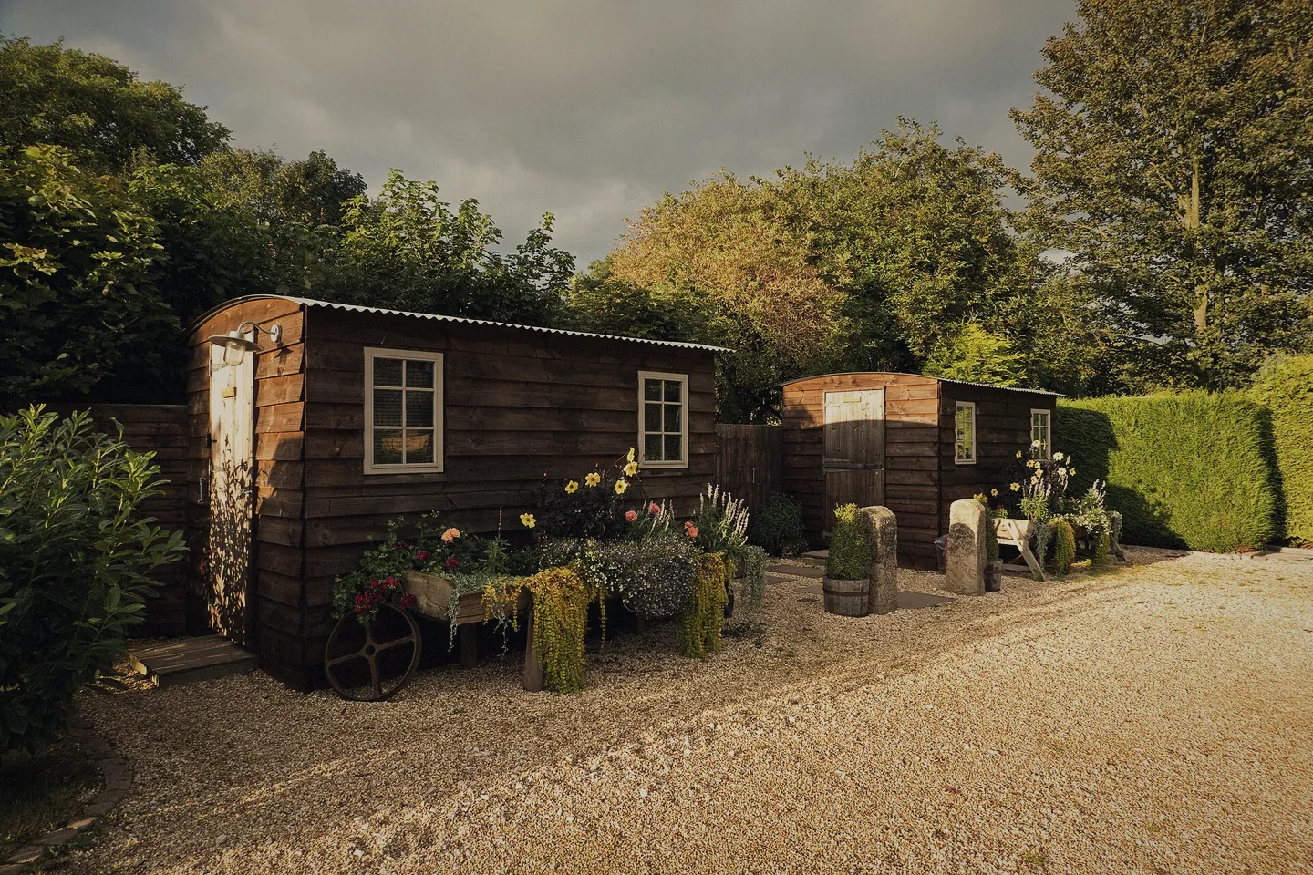 A timber shower and toilet hut on gravel with flowers in pots outside