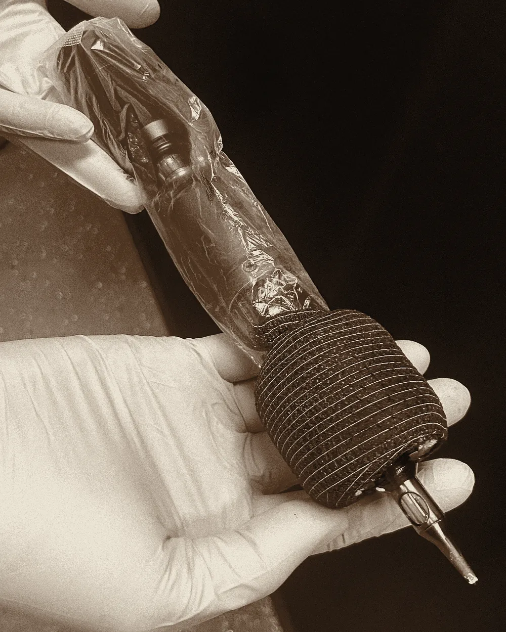 A gloved hand holding a rotary tattoo machine sleeved in barrier film