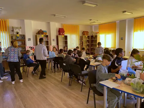 Sala de lectură cu rafturi galbene și perdele portocalii, elevi la mese