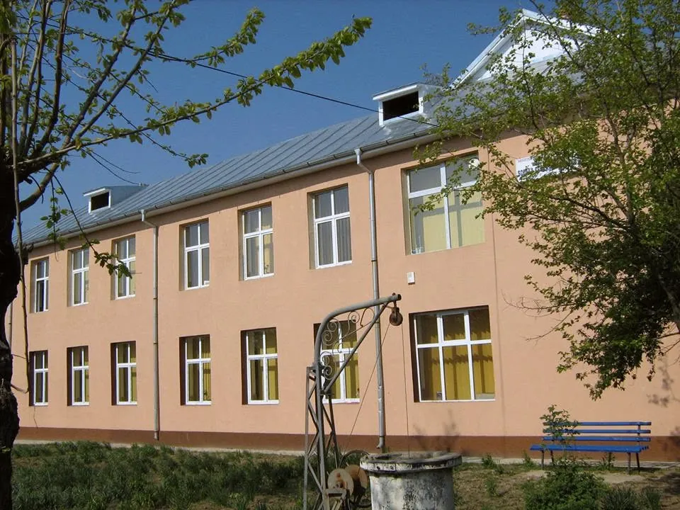Clădirea cu etaj a școlii, cu fațada de culoarea piersicii, fântâna din față și o bancă albastră sub un copac