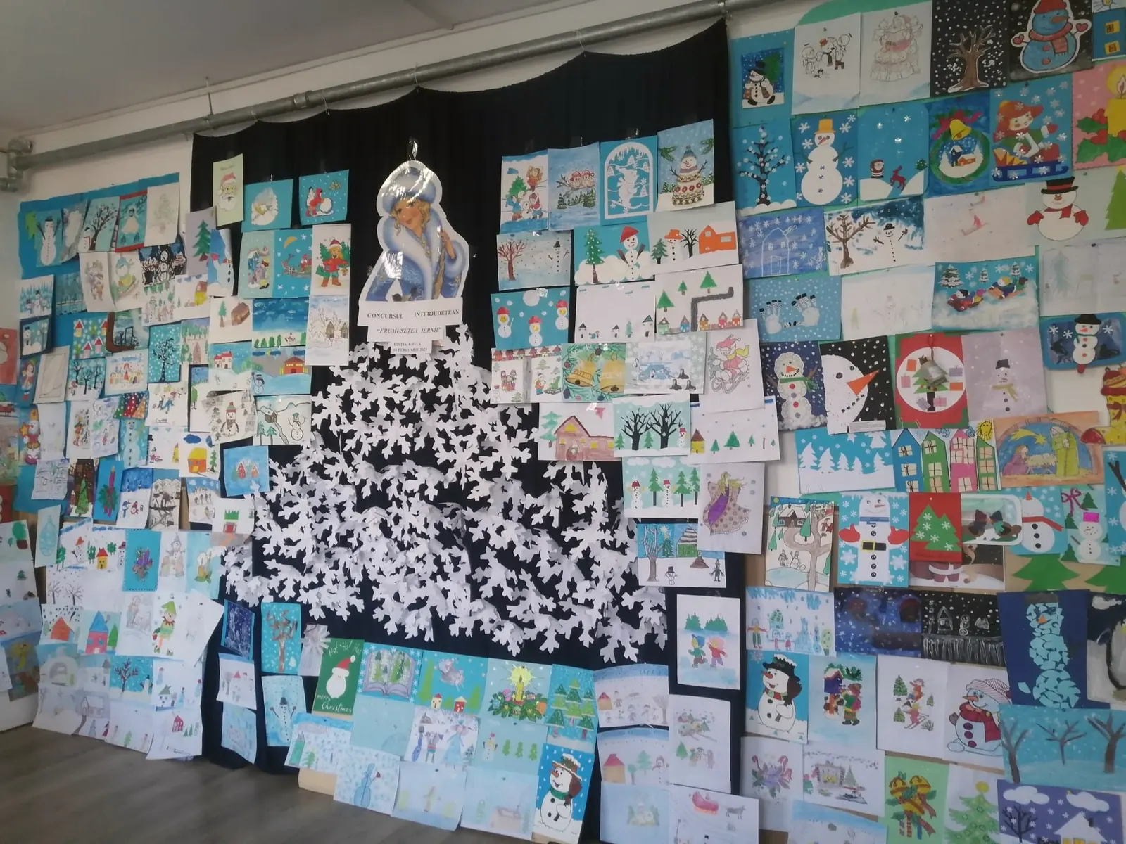 Un perete întreg acoperit de sute de desene de iarnă ale copiilor, cu un brad din fulgi de hârtie albi în mijloc