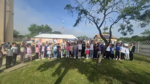 Elevi aliniați în curtea școlii, cu clădirea cu un nivel în fundal; „Micii gardieni ai curățeniei”
