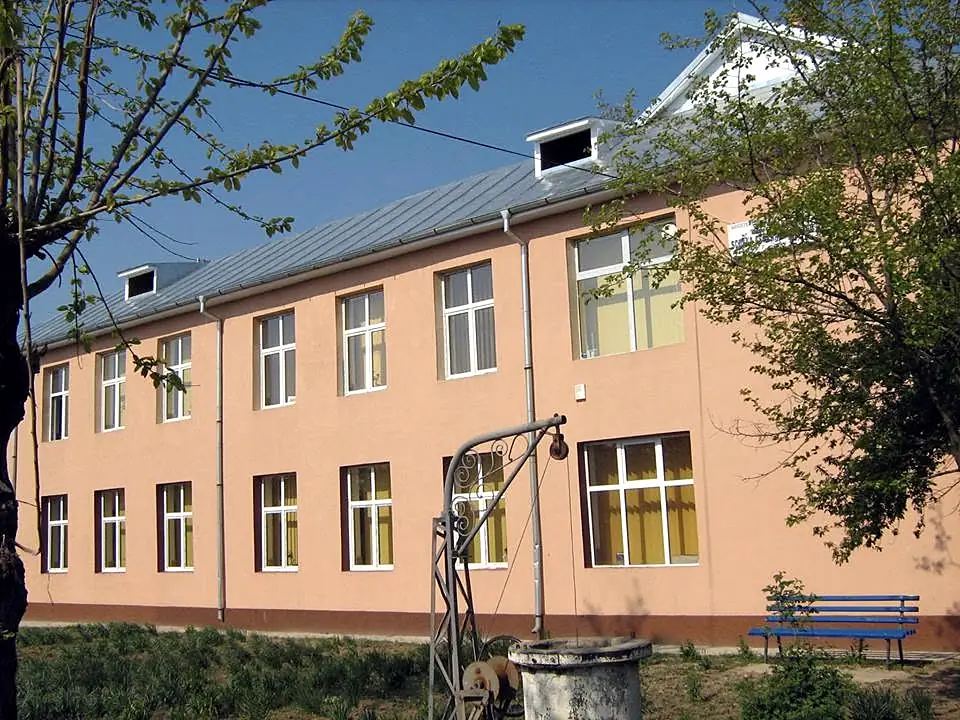 Clădirea școlii din Braneț, cu etaj, primăvara: fântâna din curte și banca albastră din fața ei